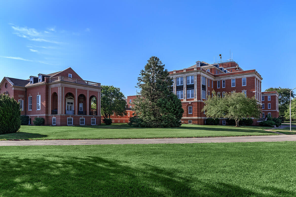 outside grassy area with 2 brick buildings in the background a road going between 2 of the grassy areas and various trees and bushes scattered throughout